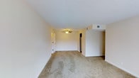 This single-floor unit at Partridge Meadows Apartments offers a straightforward, functional layout designed for comfortable everyday living. The main living area is generously proportioned, providing ample room for a variety of furniture arrangements. Neutral beige carpeting covers the floor, contributing to a warm, understated aesthetic that pairs well with the cream-colored walls throughout. A ceiling fan with a built-in light fixture is centrally positioned, ensuring both air circulation and ambient lighting in the space. HVAC vents are integrated into the ceiling, supporting efficient climate control year-round. White baseboards frame the perimeter of the room, adding a clean, finished look. A doorway visible along one wall leads to additional areas of the unit, while a glimpse of what appears to be a kitchen or utility space is accessible toward the rear. The layout is open and unobstructed, making the living area feel spacious and adaptable. The overall ambiance is clean, bright, and move-in ready, offering a neutral canvas well-suited for personalization.