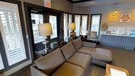 This single-floor space exudes a polished, contemporary aesthetic anchored by a thoughtfully designed lounge area. The room is furnished with a generously sized sectional sofa in a warm neutral tone, providing a comfortable and inviting focal point. Flanking the seating area are elegant silver table lamps that add a refined, decorative touch alongside carefully curated accent pieces and storage boxes arranged on a sleek console table.

The space benefits from an abundance of natural light, courtesy of multiple windows dressed with classic white plantation shutters set against bold, dark-framed surrounds that create a striking visual contrast. This interplay of light and dark tones gives the interior a sophisticated character. A cozy sitting nook near the windows features a round side table paired with an upholstered accent chair, offering an ideal spot for quiet conversation or reading.

Decorative elements such as a textured sunburst wall accent and tasteful greenery contribute to the overall warmth of the environment. The neutral color palette — encompassing soft grays, warm taupes, and rich dark trim — ties the space together cohesively. The combination of carefully selected furnishings, layered lighting, and architectural detailing makes this a welcoming and functionally versatile space suited for both relaxation and social gathering.