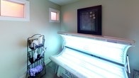 This single-room tanning space is thoughtfully arranged to provide a focused, functional environment for tanning sessions. The room is finished with soft sage-green walls that lend a calm, relaxing atmosphere, complemented by dark flooring that grounds the space visually. At the center of the room sits a Sunvision tanning bed, the clear focal point, illuminated by rows of UV lamps that cast a characteristic blue-toned glow throughout the space. A small window allows a touch of natural light to filter in, balancing the ambiance of the room. Wall décor includes a framed abstract artwork and decorative metal floral wall pieces flanking a small wall-mounted mirror with hooks, adding a personalized, welcoming touch. A wicker storage ottoman provides a convenient seating or staging surface for clients, while a wire rack organizer near the tanning bed keeps supplies such as sanitizing sprays and paper towels neatly accessible. A small fan is also on hand for client comfort during sessions. The room is accessed through a classic paneled white door with a brushed hardware lever handle, and informational signage is posted at the entry for client reference. The overall layout is compact yet efficiently organized, offering everything needed for a complete tanning session in one tidy, purpose-built room.