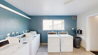 Located at 19545 SW Boones Ferry Rd in Tualatin, Oregon, this shared laundry facility is a single-floor space designed for efficient, communal use. The room is thoughtfully laid out to accommodate multiple full-sized top-loading washers and front-loading dryers arranged along the walls, providing ample capacity for residents. Light wood-style luxury vinyl flooring runs throughout, lending a clean and modern aesthetic to the utilitarian space. The interior is painted with a crisp white finish on most surfaces, while a bold teal accent wall adds a touch of character and visual interest. Natural light filters in through a centrally positioned window, offering views of the surrounding greenery and keeping the space feeling bright and airy. A ceiling fan provides additional ventilation and comfort during use. Safety is clearly a priority, with a mounted fire extinguisher and hand sanitizer dispenser visible near the entrance. A wall-mounted heater ensures year-round comfort. The space also includes an adjoining room accessible through an interior doorway, extending its functional footprint. Posted laundry room guidelines and informational signage help ensure orderly, considerate use of the facility. Overall, this laundry room is clean, well-maintained, and practical, serving as a reliable shared amenity.