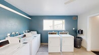 Located at 19545 SW Boones Ferry Rd in Tualatin, Oregon, this shared laundry facility is a single-floor space designed for efficient, communal use. The room is thoughtfully laid out to accommodate multiple full-sized top-loading washers and front-loading dryers arranged along the walls, providing ample capacity for residents. Light wood-style luxury vinyl flooring runs throughout, lending a clean and modern aesthetic to the utilitarian space. The interior is painted with a crisp white finish on most surfaces, while a bold teal accent wall adds a touch of character and visual interest. Natural light filters in through a centrally positioned window, offering views of the surrounding greenery and keeping the space feeling bright and airy. A ceiling fan provides additional ventilation and comfort during use. Safety is clearly a priority, with a mounted fire extinguisher and hand sanitizer dispenser visible near the entrance. A wall-mounted heater ensures year-round comfort. The space also includes an adjoining room accessible through an interior doorway, extending its functional footprint. Posted laundry room guidelines and informational signage help ensure orderly, considerate use of the facility. Overall, this laundry room is clean, well-maintained, and practical, serving as a reliable shared amenity.