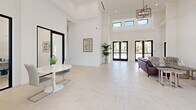 Located at 1414 SW 17th Pl in Cape Coral, Florida, this single-floor space presents a striking contemporary design defined by soaring double-height ceilings and an all-white color palette that maximizes brightness and openness. Large-format light-toned tile flooring runs throughout, creating a seamless, cohesive aesthetic across every area.

The main gathering space is grand and airy, anchored by elegant geometric chandelier fixtures suspended from the towering ceiling. Clerestory windows positioned high on the walls invite additional natural light from above, while floor-to-ceiling black-framed glass doors and windows offer views of the outdoor pool and surrounding landscape, beautifully connecting the interior to the Florida environment outside.

The lounge area features a curated arrangement of seating — including a leather sofa, patterned accent chairs, and side tables — centered around a wall-mounted flat-screen television, creating a comfortable and functional gathering hub. An adjacent conference or meeting room, enclosed by sleek black-framed glass partition doors, provides a private yet visually open space furnished with a round table and modern chairs, ideal for smaller group discussions.

Tasteful artwork and lush tropical plants are thoughtfully placed throughout, adding warmth and a Florida-inspired character to the otherwise crisp, minimalist interior. The overall ambiance is clean, sophisticated, and welcoming.