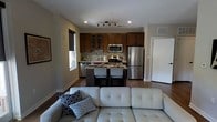 This single-floor space showcases an open-concept layout that seamlessly connects the living, dining, and kitchen areas into one cohesive environment. The kitchen is well-appointed with rich, dark wood cabinetry, stainless steel appliances including a gas range, built-in microwave, dishwasher, and a full-size refrigerator. A mosaic tile backsplash adds a decorative touch, while under-cabinet lighting enhances the workspace. A compact kitchen island with seating for two provides a casual dining option and serves as a natural divider between the kitchen and living areas. Track lighting mounted on the ceiling ensures ample illumination throughout. The living area features a spacious light gray sectional sofa accented with decorative throw pillows, creating a relaxed yet stylish atmosphere. Dark hardwood flooring runs throughout, adding warmth and continuity to the space. Neutral-toned walls complement the darker cabinetry and flooring, while a window allows natural light to filter in. The overall ambiance is modern, practical, and inviting, making efficient use of the available space.