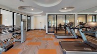 This fitness center is thoughtfully designed to support a comprehensive workout experience across two distinct zones. The main cardio and strength area is anchored by Cybex-branded machines, including treadmills, a cable/resistance system, and a recumbent stepper, all arranged to allow comfortable movement between equipment. A distinctive patterned carpet in neutral tones with bold orange accents defines the floor throughout, adding a modern, energetic aesthetic. The ceiling features a series of circular recessed coffers with integrated recessed lighting, lending architectural interest and an open, airy feel to the space. Large black-framed glass doors and floor-to-ceiling windows line one wall, inviting abundant natural light and offering views of a landscaped outdoor area. A dedicated free weight room is partitioned by a full wall of mirrors, creating a sense of expanded space. It is outfitted with a tiered dumbbell rack, adjustable benches, stability balls, and a resistance band station. The mirrored walls enhance visibility during strength training. Overall, the fitness center strikes a balance between functional utility and polished design, making it a well-rounded facility suitable for a wide range of workout routines.