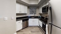 This single-floor residence offers a well-organized layout with a variety of functional spaces throughout. The living room serves as a central gathering area, finished with rich dark hardwood-style flooring that contrasts cleanly against neutral-toned walls. A fireplace with a white mantle anchors one side of the room, while a wall of windows and a sliding glass door with vertical blinds flood the space with natural light and provide direct outdoor access. A pass-through opening connects the living area to the kitchen, which features white shaker-style cabinetry, dark granite countertops, and a full suite of stainless steel appliances including a dishwasher, range, and microwave. A window above the sink adds natural light to the workspace. The bedrooms are carpeted for comfort, each equipped with a ceiling fan and natural light from windows with outdoor views. The primary bedroom includes access to a closet and an en-suite bathroom. The bathrooms feature tub-shower combinations with tiled surrounds, with one bath offering a vanity with white cabinetry and wood-look flooring. Textured ceilings are consistent throughout the space.