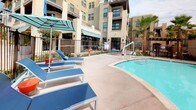 This outdoor amenity area centers around a freeform swimming pool with clear, blue water, complemented by a separate spa situated nearby. The pool deck is finished in a smooth, light-toned concrete surface that provides ample space for lounging and movement. Multiple blue chaise lounge chairs with coordinating striped umbrellas are arranged along the poolside, offering comfortable spots to relax and take in the open-air setting. An ADA-accessible pool lift is installed at the pool's edge, ensuring inclusive access for all users. The area is enclosed by a sturdy metal fence with gated entry, providing both security and a defined boundary for the space. Tall, mature palm trees are strategically placed throughout, lending a tropical, resort-like ambiance and natural shade. Landscaped planting beds with shrubs and seasonal greenery line the perimeter, softening the hardscape. The pool area is set within the interior courtyard of a multi-story residential building featuring stucco facades, large windows, and private balconies overlooking the amenity space. Views beyond the fence extend to the surrounding streetscape and neighboring structures, giving the space an urban yet relaxed character.