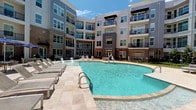 This residential unit is part of a contemporary multi-story community with a striking modern exterior, combining brick and sleek paneling accents across multiple floors. Each floor features private balconies, contributing to the building's dynamic architectural character. At the heart of the community lies a expansive resort-style courtyard pool with sparkling blue water, surrounded by a travertine deck lined with lounge chairs — ideal for relaxation and outdoor enjoyment under open skies.

Inside the unit, the open-concept living and dining area is finished with smooth hardwood-style flooring that reflects natural light streaming in through multiple windows. Ceiling fans provide comfort throughout, while neutral-toned walls offer a clean, versatile canvas. The kitchen is well-equipped, featuring rich dark wood cabinetry, grey countertops, a double-basin stainless steel sink, and a full suite of stainless steel appliances including a refrigerator, range, dishwasher, and built-in microwave.

A central hallway connects the living spaces to the private areas of the unit, with clean white panel doors leading to a bathroom and additional rooms. The hallway is finished with tile flooring that transitions seamlessly between spaces. The overall design balances practicality with modern aesthetics, offering a comfortable and cohesive living environment.