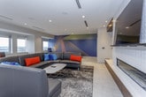 This contemporary amenity lounge is a thoughtfully designed, open-plan space that seamlessly blends social, dining, and lounge functions across a single floor. Light-toned large-format tile flooring flows throughout, lending a clean and cohesive aesthetic to every zone.

The central gathering area features a bold round dining table surrounded by vibrant orange mid-century-modern chairs, anchored beneath a striking sputnik chandelier. A wall-mounted flat-screen TV and a sleek fireplace are set against rich dark wood built-ins, while a large-scale abstract mural adds a dramatic artistic focal point. Adjacent to this, a curved sectional sofa sits atop a patterned area rug, creating an intimate lounge setting.

A dedicated dining zone offers multiple seating configurations with patterned and upholstered chairs arranged around dark tables, ideal for both casual and collaborative gatherings. Along the windowed perimeter, a banquette of dark leather seating with bold red accent pillows lines a wall of expansive windows, offering sweeping panoramic views of the surrounding cityscape and filling the space with abundant natural light.

The fully equipped kitchen features rich walnut cabinetry, white quartz countertops, a glass subway tile backsplash, stainless steel appliances including a French door refrigerator, and ample storage — providing everything needed for catering or everyday use.