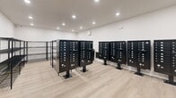 Located at 8484 Brougham Dr in Sterling Heights, Michigan, this dedicated mailroom provides a well-organized, functional space for residents to send and receive mail and packages. The room occupies a single floor and offers a comfortable amount of space to accommodate multiple users at once.

The room is anchored by several freestanding black pedestal-mounted mailbox units, each featuring numerous individually keyed compartments to serve the building's residents. The mailboxes are arranged to allow easy access and clear circulation throughout the space. Complementing these are multiple black wire shelving units positioned along the walls and centrally, providing ample surface area for package storage and retrieval.

The interior finishes are clean and modern, featuring light wood-look flooring that adds warmth to the otherwise utilitarian space. The walls are finished in a crisp white, contributing to a bright and welcoming atmosphere. Recessed ceiling lighting provides even, practical illumination throughout. A glass-paneled entry door connects the mailroom to the broader building, and an exit sign with a security camera ensures safety and compliance. A wall-mounted HVAC unit maintains a comfortable indoor environment year-round. Overall, the space is thoughtfully designed to efficiently serve the needs of a multi-unit residential community.