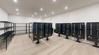 Located at 8484 Brougham Dr in Sterling Heights, Michigan, this dedicated mailroom provides a well-organized, functional space for residents to send and receive mail and packages. The room occupies a single floor and offers a comfortable amount of space to accommodate multiple users at once.

The room is anchored by several freestanding black pedestal-mounted mailbox units, each featuring numerous individually keyed compartments to serve the building's residents. The mailboxes are arranged to allow easy access and clear circulation throughout the space. Complementing these are multiple black wire shelving units positioned along the walls and centrally, providing ample surface area for package storage and retrieval.

The interior finishes are clean and modern, featuring light wood-look flooring that adds warmth to the otherwise utilitarian space. The walls are finished in a crisp white, contributing to a bright and welcoming atmosphere. Recessed ceiling lighting provides even, practical illumination throughout. A glass-paneled entry door connects the mailroom to the broader building, and an exit sign with a security camera ensures safety and compliance. A wall-mounted HVAC unit maintains a comfortable indoor environment year-round. Overall, the space is thoughtfully designed to efficiently serve the needs of a multi-unit residential community.