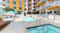 This outdoor amenity space is designed with a resort-inspired aesthetic, centered around a sparkling swimming pool and an adjacent spa. The pool deck is finished in large-format light-colored tiles, creating a clean, polished surface that reflects the surrounding architecture. Framed by the bold yellow and white exterior of the multi-story building, the courtyard setting lends the space an enclosed yet open-air feel, with natural light flooding in throughout the day.

The lounge area is well-appointed with a generous selection of adjustable chaise lounges and coordinating teal accent tables, all shaded by vibrant turquoise umbrellas. Woven circular lounge chairs with accent pillows provide additional seating options, adding a contemporary touch. Built-in hardwood benches mounted on concrete bases are positioned near lush greenery and mature trees, offering a quieter, more secluded retreat within the space.

Glass panel railings with brushed stainless steel hardware delineate different zones of the deck while maintaining visual openness. Textured bamboo wall cladding at the entry adds a warm, natural contrast to the otherwise modern palette. Raised planters with ornamental grasses and shrubs enhance the landscaping throughout, contributing to a vibrant, well-maintained outdoor environment.