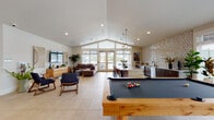 This single-story community clubhouse offers a thoughtfully designed amenity space that seamlessly blends recreation, relaxation, and social gathering. The open-plan layout is anchored by large-format light tile flooring throughout, creating a cohesive and airy atmosphere.

The main gathering area centers on a vaulted ceiling with a striking clerestory window that floods the space with natural light. French doors open to outdoor views, enhancing the sense of openness. A full-size pool table anchors the game area, complemented by a colorful prize wheel for entertainment.

A standout fireplace clad in light brick tile rises dramatically toward the ceiling, flanked by a warm wood-paneled accent wall. Built-in bench seating and a dining table create an intimate lounge nook beside it.

The kitchenette is a design highlight, featuring dark wood cabinetry, a white quartz island with bar seating, stainless steel appliances, and a bold geometric white tile backsplash illuminated by industrial cage pendant lights.

A dedicated lounge area includes a wall-mounted TV, comfortable seating, and curated décor. A glass-doored fitness room is accessible directly from the lounge, housing exercise equipment. The overall ambiance is modern, warm, and welcoming throughout.