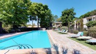 This single-story property in Pensacola offers an inviting outdoor amenity area centered around a generously sized swimming pool with crystal-clear blue water. The pool features clearly marked depth indicators and entry steps, making it accessible and user-friendly. Surrounding the pool is a well-maintained paved deck constructed from attractive brick-patterned pavers, providing ample space for lounging and entertaining. Multiple chaise lounge chairs with teal cushions are arranged along the poolside, offering comfortable spots to relax and soak in the Florida sunshine. A shaded seating area with an umbrella provides a cool retreat from the sun. The outdoor space is framed by mature, leafy trees that offer natural shade and privacy, creating a serene atmosphere. Lush greenery and landscaping throughout the area enhance the tropical ambiance. A wrought iron perimeter fence encloses the pool area, adding both a safety feature and a classic aesthetic element. The backdrop reveals the building's architectural presence, nestled among the verdant foliage under a clear blue sky, embodying the relaxed, sun-soaked lifestyle characteristic of coastal Pensacola.