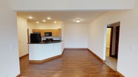 This single-floor space showcases an open-concept layout that seamlessly connects the kitchen and living areas, creating a cohesive and functional environment. The kitchen is equipped with light honey-toned wood cabinetry that provides ample storage, complemented by dark countertops that offer a striking contrast. Black appliances, including a refrigerator and microwave, are integrated throughout, lending a sleek, uniform look. A curved breakfast bar serves as a natural divider between the kitchen and the adjacent living space, making it ideal for casual dining or entertaining.

Recessed lighting in the kitchen ensures bright, even illumination, while a flush-mount ceiling fixture extends light into the open living area. The warm, medium-toned hardwood-style flooring runs continuously throughout, adding warmth and visual cohesion to the space. Crisp white walls keep the interior feeling bright and open. A doorway visible toward the rear of the living area suggests additional rooms beyond, hinting at further functional space. The overall design is clean, practical, and move-in ready.