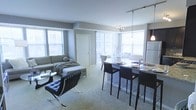 This contemporary single-floor residence showcases a thoughtfully designed open-concept layout that seamlessly connects the living, dining, and kitchen areas into one cohesive, flowing space. Large windows line the exterior walls, flooding the interior with abundant natural light and offering views of the surrounding urban landscape.

The living area is anchored by a plush sectional sofa and complemented by a classic Barcelona-style accent chair, resting atop soft wall-to-wall carpeting that adds warmth to the modern aesthetic. A glass coffee table and understated décor complete the refined yet comfortable atmosphere.

The kitchen and dining zone anchor the opposite end of the open floor plan, featuring a granite-topped breakfast bar with sleek dark bar stools — ideal for casual dining or entertaining. A pendant light and track lighting overhead provide both task illumination and ambient warmth. The kitchen is equipped with full-size appliances and dark cabinetry, offering a striking contrast against the light-toned walls.

A formal dining area sits adjacent, bathed in natural light from the expansive windows, creating an inviting setting for shared meals. The overall palette of cool grays, blacks, and whites gives the space a clean, sophisticated character throughout.