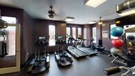 Located at 425 Wilcox St in Charlotte's South End neighborhood, this fitness center offers a well-equipped, single-floor workout facility designed for comprehensive training. The space is finished with deep purple-toned walls and durable rubber flooring, creating a modern, energetic atmosphere throughout.

The cardio area features multiple treadmills and elliptical machines, each equipped with individual digital screens, positioned along a windowed wall that allows natural light and outdoor views of the surrounding area. Ceiling fans provide additional air circulation throughout the space.

The strength training section includes a cable machine system, a power rack with weight plates, a full dumbbell rack, and various resistance machines. Functional training accessories such as medicine balls, stability balls, and exercise mats are also available and neatly stored.

Exposed metallic HVAC ductwork runs across the ceiling, contributing to an industrial aesthetic that complements the overall design. The facility connects through glass-paneled doors to adjacent common areas, enhancing accessibility. Recessed and flush-mounted ceiling lighting ensures the entire space remains well illuminated, supporting a practical and motivating workout environment.