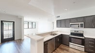 This single-floor unit presents a clean, contemporary design characterized by an open layout, wood-style flooring, and a crisp white color palette throughout. The kitchen serves as a central highlight, featuring dark espresso-toned cabinetry with brushed nickel hardware, light quartz countertops, and a full suite of stainless steel appliances including a refrigerator, electric range, microwave, and dishwasher. A peninsula island extends the counter space and provides a natural divide between the kitchen and the adjacent living area, which is bathed in natural light through black-framed windows. Recessed lighting supplements the natural light across all main areas. The bedrooms are generously proportioned, each featuring the same wood-style flooring, white walls, and black-framed windows that frame outdoor views and allow ample sunlight to fill the rooms. Multiple doors in the primary bedroom suggest access to a closet and an en-suite bathroom. The bathroom is outfitted with a dual-sink white vanity, a wide wall-to-wall mirror, and modern chrome fixtures, creating a bright and functional space. The overall ambiance is modern, light-filled, and move-in ready.