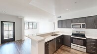 This single-floor unit presents a clean, contemporary design characterized by an open layout, wood-style flooring, and a crisp white color palette throughout. The kitchen serves as a central highlight, featuring dark espresso-toned cabinetry with brushed nickel hardware, light quartz countertops, and a full suite of stainless steel appliances including a refrigerator, electric range, microwave, and dishwasher. A peninsula island extends the counter space and provides a natural divide between the kitchen and the adjacent living area, which is bathed in natural light through black-framed windows. Recessed lighting supplements the natural light across all main areas. The bedrooms are generously proportioned, each featuring the same wood-style flooring, white walls, and black-framed windows that frame outdoor views and allow ample sunlight to fill the rooms. Multiple doors in the primary bedroom suggest access to a closet and an en-suite bathroom. The bathroom is outfitted with a dual-sink white vanity, a wide wall-to-wall mirror, and modern chrome fixtures, creating a bright and functional space. The overall ambiance is modern, light-filled, and move-in ready.