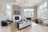 Located at 10571 Colorado Blvd in Thornton, Colorado, this single-floor space combines a kitchen and living room into one cohesive, open-plan layout designed with modern sensibilities. The living area commands attention with its dramatically vaulted ceiling, anchored by a sleek gas fireplace set against a textured accent wall with a natural wood mantle. A striking sputnik chandelier hangs overhead, adding an architectural focal point. Light-toned wood-look flooring runs throughout, contributing to the bright, airy atmosphere. An abundance of oversized windows along multiple walls floods the space with natural light and frames views of the surrounding outdoor greenery. The living area is thoughtfully arranged with comfortable seating, a stylish media console, and coordinating accent tables, offering both functionality and visual appeal. Adjacent to the living space, the kitchen features crisp white cabinetry with matte black hardware, a stainless steel sink, and textured tile backsplash extending to the ceiling. It is equipped with a compact refrigerator and microwave, making it well-suited for a lounge or amenity setting. A cozy seating nook near the kitchen window provides an intimate corner bathed in natural light. The overall ambiance is clean, contemporary, and welcoming.