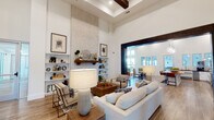 This single-floor community space blends modern farmhouse design with sophisticated, welcoming interiors. The main living area commands attention with dramatically soaring ceilings accented by dark exposed wood beams and a striking floor-to-ceiling stone fireplace surround with a rustic wood mantel, flanked by built-in shelving. Wood-look tile flooring flows throughout, lending warmth and continuity across each zone.

The open-plan layout connects multiple seating and gathering areas through bold, dark-framed architectural passageways. Neutral-toned sofas, upholstered accent chairs, and a mix of natural wood coffee and side tables create inviting conversation areas. Shiplap wall paneling is a recurring design element, contributing to the space's cohesive farmhouse aesthetic.

A dedicated lounge area features large floor-to-ceiling windows and glass doors that open to an outdoor courtyard, flooding the interior with abundant natural light and views of the exterior amenity areas. A communal dining zone centers around a substantial live-edge wood table beneath globe pendant lights.

The kitchenette area offers dark countertops, white cabinetry, open wood floating shelves, a beverage refrigerator, and a wall-mounted television set against a textured brick accent wall — a well-equipped space for social gatherings and events.