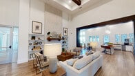 This single-floor community space blends modern farmhouse design with sophisticated, welcoming interiors. The main living area commands attention with dramatically soaring ceilings accented by dark exposed wood beams and a striking floor-to-ceiling stone fireplace surround with a rustic wood mantel, flanked by built-in shelving. Wood-look tile flooring flows throughout, lending warmth and continuity across each zone.

The open-plan layout connects multiple seating and gathering areas through bold, dark-framed architectural passageways. Neutral-toned sofas, upholstered accent chairs, and a mix of natural wood coffee and side tables create inviting conversation areas. Shiplap wall paneling is a recurring design element, contributing to the space's cohesive farmhouse aesthetic.

A dedicated lounge area features large floor-to-ceiling windows and glass doors that open to an outdoor courtyard, flooding the interior with abundant natural light and views of the exterior amenity areas. A communal dining zone centers around a substantial live-edge wood table beneath globe pendant lights.

The kitchenette area offers dark countertops, white cabinetry, open wood floating shelves, a beverage refrigerator, and a wall-mounted television set against a textured brick accent wall — a well-equipped space for social gatherings and events.