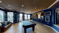 This single-story clubroom in Spring, TX is designed as a well-appointed social and recreational hub. Bold navy blue walls with white crown molding establish a strong visual identity throughout the space, complemented by warm-toned tile flooring and a dark ceiling with recessed lighting. The main gathering area centers around a full-sized pool table, providing a focal point for recreation and entertainment.

A fully equipped kitchen anchors one end of the clubroom, featuring warm wood cabinetry, granite countertops, stainless steel appliances including a refrigerator and range, and pendant lighting that adds a modern touch. Adjacent to the kitchen, a bar-height counter with metal barstools offers additional seating, making the space well-suited for social gatherings.

A comfortable lounge area lines the windowed perimeter of the room, furnished with rich brown leather armchairs and a sofa accented with decorative pillows. Oversized windows fitted with blinds and dark drape panels allow ample natural light while offering views of the surrounding outdoor greenery. A small dining nook with a round table and modern chairs provides an additional casual seating option. The overall ambiance is energetic yet comfortable, balancing recreational functionality with relaxed social appeal.