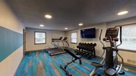 Located at 7025 Bronner Cir in Louisville, Kentucky, this well-equipped fitness center offers a comprehensive workout environment on a single floor. The space is immediately distinguished by its eye-catching multi-tone carpet flooring in rich shades of teal, blue, and warm wood-toned hues, adding a modern and energetic character to the room.

The cardio section features multiple treadmills positioned near windows with blinds that allow natural light to filter in, creating a bright and motivating atmosphere. An elliptical trainer and stationary cycling bikes round out the cardio offerings, catering to a variety of fitness preferences. A full rack of dumbbells in graduated weights is neatly organized along one wall, accompanied by an adjustable workout bench for strength training exercises.

A wall-mounted flat-screen television is prominently positioned for easy viewing from multiple workout stations, enhancing the exercise experience. Recessed lighting throughout the textured ceiling ensures the space is well-lit at all times. Large sliding glass doors connect the fitness center to an adjacent lounge or common area, promoting a sense of openness and easy access between spaces. The neutral gray walls provide a clean, focused backdrop, while wall art adds a subtle decorative touch. Overall, the fitness center presents a functional, modern, and welcoming environment for daily exercise.