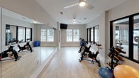 This yoga and multi-purpose fitness room is a well-appointed studio space designed to support a variety of exercise disciplines. The room features light-toned hardwood flooring throughout, creating a clean and energizing environment conducive to both high-intensity and mindful workout sessions. Full-length mirrors line one wall, allowing users to monitor their form during yoga, stretching, or cycling sessions, while also enhancing the sense of openness within the space.

The studio is equipped with several spinning bikes positioned along one side of the room, accompanied by an assortment of stability and exercise balls in various sizes. Ceiling fans provide consistent air circulation, contributing to a comfortable workout environment. Natural light enters through a set of windows on one end of the room, adding brightness and a connection to the outdoors.

Large glass-paneled windows along an interior wall offer direct sightlines into an adjacent, fully equipped fitness center featuring cardio machines and strength training equipment. A glass-and-black-framed door provides access between the two spaces. The overall design is clean and modern, with neutral gray walls and white trim that maintain a focused, professional fitness atmosphere throughout.