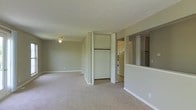 This single-floor space offers a straightforward, functional layout designed for comfortable everyday living. The main living area is generously proportioned, featuring neutral-toned carpet flooring and soft beige walls that create a calm, understated ambiance. A ceiling fan is centrally mounted, providing both lighting and air circulation. A built-in closet with bifold doors is conveniently positioned within the living space, offering practical storage with a hanging rod and shelf visible inside. To one side, a half-wall with a pass-through opening connects the living area to the kitchen, promoting an open feel while maintaining a degree of separation between the spaces. Built-in shelving is visible on the adjacent wall near the pass-through, adding additional storage or display options. The kitchen, partially visible through the opening, features white cabinetry and white appliances, maintaining a clean and consistent aesthetic throughout. White baseboards run along the perimeter of the living area, providing a crisp contrast to the neutral carpet and walls. Overall, the space presents a clean, move-in-ready environment with a simple, practical design.