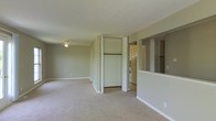 This single-floor space offers a straightforward, functional layout designed for comfortable everyday living. The main living area is generously proportioned, featuring neutral-toned carpet flooring and soft beige walls that create a calm, understated ambiance. A ceiling fan is centrally mounted, providing both lighting and air circulation. A built-in closet with bifold doors is conveniently positioned within the living space, offering practical storage with a hanging rod and shelf visible inside. To one side, a half-wall with a pass-through opening connects the living area to the kitchen, promoting an open feel while maintaining a degree of separation between the spaces. Built-in shelving is visible on the adjacent wall near the pass-through, adding additional storage or display options. The kitchen, partially visible through the opening, features white cabinetry and white appliances, maintaining a clean and consistent aesthetic throughout. White baseboards run along the perimeter of the living area, providing a crisp contrast to the neutral carpet and walls. Overall, the space presents a clean, move-in-ready environment with a simple, practical design.