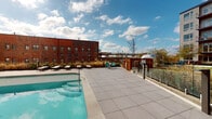 Located at 2525 E Main St in Richmond, Virginia, this amenity space combines contemporary interior design with impressive outdoor features. The interior lounge area is finished with a calming two-tone color scheme — soft white upper walls transitioning into a muted blue-gray wainscoting — complemented by patterned carpet flooring in warm earth tones with teal accents. The drop-panel ceiling is outfitted with recessed and flush-mount lighting, creating a well-lit, comfortable environment. Modern lounge seating, including sleek upholstered chairs and accent tables, is arranged throughout the space, offering a relaxed and welcoming atmosphere. Built-in cabinetry and counter space along the walls provide functional utility within the lounge. Large glass double doors open directly to an outdoor walkway, flooding the interior with natural light and offering views of the surrounding residential community and landscaped greenery. Outside, a rooftop pool deck features a sparkling pool, lounge chairs, and seating areas set against a backdrop of Richmond's urban skyline, including the characteristic red brick architecture of the area. Glass railings along the rooftop perimeter ensure unobstructed views of the cityscape under open skies.