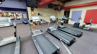 This fitness center combines rustic architectural character with modern exercise amenities in a spacious, single-floor layout. Exposed heavy timber posts and beam ceilings give the space a distinctive industrial-lodge aesthetic, complemented by bold accent colors including chartreuse green, purple, and red throughout the interior walls and ceiling ductwork.

The cardio zone is well-appointed with multiple SportsArt treadmills, each equipped with individual digital displays, arranged in rows across durable, multi-tone carpeted flooring. Overhead, wall-mounted flat-screen televisions provide entertainment for users during workouts, while large industrial ceiling fans ensure air circulation throughout the space.

A dedicated open workout area features mirrored walls, purple accent paint, and ample floor space suitable for functional training, stretching, or group exercise. Kettlebells and other freestanding equipment are available in this zone. Interior windows along a curved partition wall allow visual connectivity between areas while maintaining distinct workout zones.

Natural light enters through windows and glass double doors along the perimeter, offering views of the surrounding brick exterior. The facility is well-lit with linear fluorescent fixtures mounted to the exposed beam ceiling, creating a bright and energetic training environment throughout.