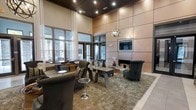 This single-floor space exudes a refined, contemporary aesthetic characterized by its dramatic double-height ceilings and expansive wall paneling finished in warm, brushed copper tones. Recessed lighting embedded in the dark ceiling grid provides ambient illumination, complemented by a striking spherical chandelier that serves as a focal point above the central seating area. A built-in fireplace framed within the feature wall adds warmth and sophistication, with a mounted flat-screen display positioned above it. The lounge area is anchored by a sleek, low-profile coffee table surrounded by plush, dark upholstered armchairs and sofas arranged atop a textured area rug, creating an inviting conversation space. Floor-to-ceiling windows and glass-paneled doors along one wall flood the interior with natural light while offering views of the exterior surroundings. Sheer white drapery panels soften the window wall, adding an elegant contrast to the bold architectural elements. Light-toned tile flooring borders the carpeted lounge zone, enhancing the sense of spaciousness. Overall, the space conveys a polished, hospitality-inspired ambiance suited for relaxed gatherings.