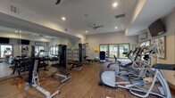Located at Cameron Woods in Apex, NC, this well-appointed fitness center offers a comprehensive workout environment on a single floor. The space features warm hardwood-style flooring throughout, elevated ceilings with ceiling fans, and recessed lighting that create a bright and open atmosphere. Large windows along multiple walls invite abundant natural light and provide views of the surrounding greenery, enhancing the workout experience.

The cardio zone is equipped with Precor treadmills, elliptical trainers, and other aerobic machines positioned along the windowed walls, each accompanied by wall-mounted flat-screen televisions for entertainment during workouts. The strength training area includes a multi-station cable machine, adjustable weight benches, a full dumbbell rack, and kettlebells of varying sizes, accommodating a wide range of resistance training routines. Stability and medicine balls are available for functional fitness exercises.

Floor-to-ceiling mirrors line one wall, visually expanding the space and allowing users to monitor their form. A water fountain and built-in shelving provide added convenience. The neutral color palette of soft gray and white walls, combined with the warm flooring and ample natural light, creates an inviting and energizing environment well-suited for all fitness levels.
