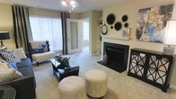 This single-story space centers around a welcoming living area designed with both comfort and style in mind. The room features a classic fireplace with a crisp white mantle, serving as the focal point of the space, accented by decorative black wall art and metallic accessories displayed above. Built-in shelving flanks the fireplace, offering functional storage and display space. Neutral, warm-toned walls complement the plush carpeted flooring, creating an inviting atmosphere throughout. Large windows dressed with horizontal blinds and dark curtain panels allow generous natural light to fill the room while maintaining privacy. A modern chandelier provides additional overhead lighting, adding a touch of elegance to the space. The living area is thoughtfully arranged with a sofa, a glass-top coffee table, and a pair of round upholstered ottomans, balancing practicality with aesthetic appeal. French-style interior doors with glass panels lead to an adjacent room, enhancing the open and airy feel of the layout. The overall ambiance is calm and sophisticated, with a cohesive neutral palette accented by bold decorative elements.