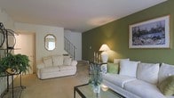 This two-floor residence opens into a well-appointed living room that immediately establishes a warm and inviting ambiance. A striking olive green accent wall serves as the room's focal point, providing rich contrast against the surrounding neutral white walls and light-toned carpeted flooring. The space is furnished with two cream-colored sofas arranged around a glass-topped coffee table, creating a comfortable and elegant seating area. Decorative touches include a candelabra side table with a classic white table lamp, framed wall art, and a gold-framed octagonal mirror that adds a traditional charm to the space. A glass vase with blue florals on the coffee table introduces a subtle pop of color. An iron-railed staircase with carpeted treads connects the main level to the upper floor, adding an architectural element that enhances the overall character of the home. The layout feels open and cohesive, with thoughtful design details throughout that contribute to a refined yet comfortable living environment suited for both relaxed daily living and entertaining.