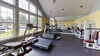 Located at 70 Greenway Sq in Dover, Delaware, this fitness center offers a well-appointed, single-floor workout facility designed to support a comprehensive exercise routine. The space is characterized by soaring vaulted ceilings that create an open, airy atmosphere, while large multi-pane windows along one wall flood the room with natural light and offer views of the surrounding greenery outside.

The interior features soft yellow walls accented by a gray wainscoting band, giving the space a clean and energizing aesthetic. Durable rubber flooring covers the entire workout area, providing both safety and comfort underfoot. A full-length wall mirror runs along one side of the room, enhancing the sense of space and allowing users to monitor their form during exercise.

The facility is equipped with a variety of SportsArt and PowerLine cardio machines, including multiple treadmills and stationary bikes, as well as a rowing machine. A selection of strength training machines with adjustable seats and cable systems is arranged throughout the space, offering options for targeting multiple muscle groups. French doors at the far end of the room provide an additional entry point and contribute to the bright, welcoming environment. The overall layout is thoughtfully organized to allow for comfortable movement between stations.