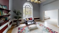 Located at 5538 S Hyde Park Blvd in Chicago's historic Hyde Park neighborhood, this single-floor space combines classical architectural character with functional, modern use. The interior is defined by impressively tall ceilings supported by exposed white beamed soffits, creating an open and airy atmosphere throughout the main area.

The flooring is a standout feature — polished terrazzo in a light speckled finish runs throughout, accented by a distinctive mustard-gold inlay stripe that guides movement through the space. Gray-painted walls are complemented by rich burgundy architectural details, including bold arched niches that serve as decorative focal points. One niche houses built-in shelving stocked with books, adding a welcoming, community-oriented character.

A dramatic arched window dominates one end of the main hall, flooding the space with natural light and offering views of the surrounding streetscape. A seating area with a colorful area rug anchors this end of the room. Elsewhere, a windowed interior partition separates an administrative or office area, and a long corridor connects to additional rooms. Industrial-style pendant lighting and a combination of lounge and work furniture complete the functional layout.