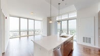 Located at Harbor Point in Stamford, Connecticut, this generously sized 2-bedroom, 1-bathroom residence offers a sophisticated modern design with an abundance of natural light. The open-concept kitchen and living area is a standout, featuring soaring ceilings, expansive floor-to-ceiling windows, and rich hardwood flooring. The kitchen is well-appointed with a large center island topped with bright white countertops, warm wood cabinetry extending to the ceiling, stainless steel appliances including a gas range, microwave, and refrigerator, and pendant lighting that adds a refined touch. The living area flows seamlessly from the kitchen, bathed in light from wraparound windows that frame sweeping panoramic views of the Stamford skyline and waterway below. Both bedrooms are carpeted and generously proportioned, with the primary bedroom offering impressive floor-to-ceiling windows showcasing direct harbor and city views. The bathroom is cleanly finished with wood-toned tile flooring, a combination tub and shower with white tile surround, a wooden vanity with white countertop, and warm wall tones. A connecting hallway ties the layout together efficiently. This residence delivers an elevated urban living experience with striking views at every turn.