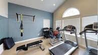 This dedicated fitness room offers a comprehensive workout environment designed for versatile training. The space is characterized by vaulted ceilings that create an open, airy feel, complemented by neutral carpeted flooring and a two-tone color scheme — a calming slate-blue accent wall contrasting with warm beige on the remaining walls.

Natural light filters in through multiple windows fitted with white horizontal blinds, including a prominent arched transom window that serves as a distinctive architectural focal point. The room is well-equipped with a full-size treadmill, a stationary exercise bike, and an adjustable weight bench for strength training. A wall-mounted pull-up bar with TRX suspension straps provides functional training options, while a folding exercise mat stored nearby adds versatility. A tiered dumbbell rack holds a range of free weights, and medicine balls round out the available equipment.

A recessed ceiling light provides additional illumination, and a wall-mounted paper towel and sanitizer dispenser reflects thoughtful, practical detailing. The entry area features framed fitness center rules, contributing to the organized and purposeful character of the space.