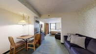 Located in Port Clinton, Ohio—known as the "Walleye Capital of the World"—Unit 205 at 2039 E Harbor Rd is a efficiently designed one-bedroom, one-bathroom suite situated on the second floor. The unit is organized into distinct functional zones despite its compact footprint. The main living area features carpet flooring, neutral cream-toned walls, and a drop ceiling, creating a straightforward and practical atmosphere. This space accommodates a seating area with a leather recliner and a sofa, alongside a small dining nook furnished with a bistro-style table and two wooden chairs. A dresser with mirror and pendant lamp add to the utilitarian character of the living space. The bedroom is accessed through an open doorway and features a window that allows natural light to enter, carpeted flooring, and room for a full-sized bed with a wooden headboard and nightstand. The bathroom is equipped with tile flooring, a shower-tub combination with a curtain, toilet, towel bar, and accessibility grab bars, reflecting practical design considerations. Overall, the unit presents a no-frills, functional layout suitable for straightforward accommodations near the Lake Erie waterfront.