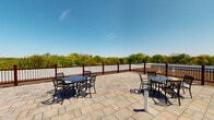 This rooftop terrace is a well-appointed outdoor space designed for relaxation and social gatherings, offering an elevated vantage point with panoramic views of the surrounding landscape. The floor is finished with large-format stone pavers in a natural, earthy tone, lending a clean and durable surface that complements the open-air setting.

The terrace is enclosed by a distinctive railing system featuring dark metal balusters set between rich, dark red posts, providing both safety and a polished aesthetic. The space is divided into functional zones: a seating and dining area furnished with ornate cast-iron tables and chairs, ideal for outdoor meals or conversation, and a sunbathing area outfitted with a pair of reclining lounge chairs positioned to take full advantage of the open sky.

A green roof section is visible from the terrace, reflecting an eco-conscious design approach. Access to the terrace is provided through a bold blue door set into a white lap-sided structure, adding a pop of color to the roofline. Solar-style bollard posts are stationed across the deck, suggesting ambient lighting for evening use. The sweeping treetop and neighborhood views, combined with the generous open space, create a bright and inviting outdoor retreat.