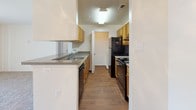 This single-floor unit is designed with a straightforward, functional layout that maximizes the use of available space across its rooms. The living area is finished with neutral-toned carpet and features a large sliding glass door fitted with vertical blinds, allowing abundant natural light to fill the room while providing direct access to an outdoor area with exterior views.

The galley-style kitchen is well-equipped with warm wood-toned cabinetry, laminate countertops, and hardwood-style flooring. Black appliances, including a refrigerator, gas range, and dishwasher, line both sides of the corridor-style space, complemented by a double-basin sink and overhead lighting.

The bedrooms continue the neutral carpet flooring and light-colored walls. One bedroom features a window with blinds that invites natural light, while another is a more enclosed, quieter space — both offering clean, blank-slate environments.

The bathroom includes a full-size soaking tub and shower combination with white tile surround and chrome fixtures. A wood-toned vanity with a granite-style countertop, large wall mirror, and multi-bulb lighting fixture complete the space. The bathroom floor features hardwood-style planking, adding warmth to the otherwise crisp, white interior.