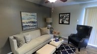 This single-floor space exudes a refined, contemporary aesthetic from the moment you step inside. The living area serves as the centerpiece, showcasing a cohesive design palette of soft grays, deep navy, and warm metallic accents that combine to create an inviting yet polished atmosphere. A plush, neutral-toned sofa anchors the seating area, complemented by upholstered ottomans that double as a coffee surface and a classic navy blue geometric-patterned area rug that grounds the space with bold contrast. A sleek black accent chair adds a sophisticated edge to the arrangement. The walls are adorned with two striking framed art pieces — one featuring an abstract oceanic landscape in blues and greens, and another displaying a floral composition — lending the room a gallery-like quality. A textured ceiling with a ceiling fan overhead adds architectural character, while pendant lighting provides a warm, ambient glow. A silver table lamp atop a side table contributes additional layers of light and elegance. Hardwood-style flooring runs throughout, offering a clean and durable foundation that ties the design together. The overall ambiance is welcoming and thoughtfully curated, reflecting a balance of comfort and contemporary style.