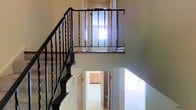 This two-story space is defined by its central staircase, which serves as a focal architectural element connecting both floors. The staircase features dark wood treads paired with ornate wrought iron balusters, adding a classic, elegant detail to the interior. A wooden handrail runs along the length of the stairs, complementing the dark metal work beneath it.

At the upper level, an open landing overlooks the lower floor, reinforcing the vertical openness of the space. A window positioned at the top of the stairwell allows natural light to filter down through both levels, brightening the surrounding neutral-toned walls.

The lower level reveals a hallway that connects to multiple areas, including what appears to be a kitchen or cabinetry area visible in the background, as well as a bathroom or utility space. The light-colored flooring on the ground level reflects available light, contributing to a clean and open feel throughout.

Overall, the space presents a functional yet characterful layout, with the interplay of wrought iron, wood, and neutral finishes creating a cohesive and timeless aesthetic across both floors.