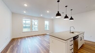 Located at 1503 Robert Weaver Ave in Austin, TX, this single-floor unit offers a clean, contemporary design with an open-concept layout that seamlessly connects the kitchen and living areas. Rich wood-look flooring runs throughout the main living spaces, creating a warm and cohesive aesthetic.

The kitchen is a standout feature, centered around a large white island with a quartz-style countertop and an integrated sink. A trio of bold matte black pendant lights hang above the island, adding a modern industrial accent. White shaker-style cabinetry lines the walls, complemented by a subway tile backsplash and stainless steel appliances, including a refrigerator, range, and microwave. Recessed lighting provides additional illumination throughout.

The open living area benefits from multiple windows that invite natural light and offer views of the surrounding neighborhood. A separate bedroom features plush carpet flooring and a ceiling fan, while an additional room with wood-look flooring and a window provides flexible use. The bathroom is finished with a glass-enclosed walk-in shower, wood-look flooring, and a white vanity with a gray countertop, maintaining the unit's consistent modern palette.