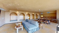 Located at 200 Mesnager St in Los Angeles, this thoughtfully designed single-floor communal space offers a warm, inviting atmosphere that blends Mediterranean-inspired architecture with modern amenities. The interior is defined by a striking row of arched wall niches lined with plywood backing and cushioned window-seat benches, creating intimate alcoves ideal for relaxing or socializing. Warm sandy-toned plaster walls and ceilings complement the large-format checkered stone tile flooring, lending the space a cohesive, earthy aesthetic throughout. A generously sized dark communal table anchors the dining area, surrounded by rustic wooden stools and illuminated by a sculptural yellow pendant light overhead. Adjacent to the dining area, a tiled wet bar or kitchenette with mosaic detailing provides functional support for gatherings. The lounge area features a plush, ruffled blue sofa paired with a rounded white side table set with a chess board, adding a playful, leisurely touch. A wall-mounted flat-screen TV completes the entertainment setup. Floor-to-ceiling glass doors open onto an outdoor terrace that captures sweeping views of the Los Angeles skyline, flooding the interior with abundant natural light. Recessed ceiling lighting ensures a well-lit environment at all hours.