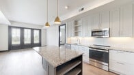 This contemporary single-floor residence showcases a thoughtfully designed open-concept layout that seamlessly blends style and functionality. The heart of the home is the expansive kitchen, which features crisp white shaker cabinetry extending to the ceiling, a classic white subway tile backsplash, and a full suite of stainless steel appliances including a range, microwave, dishwasher, and refrigerator. A central island with a contrasting gray base and a striking granite countertop provides additional workspace and storage, while a pair of elegant brass pendant lights add a warm, modern touch overhead. Wide-plank hardwood flooring flows throughout the main living areas, complementing the neutral color palette and creating a cohesive, inviting atmosphere. The living spaces are bright and airy, benefiting from abundant natural light streaming through large windows dressed with clean white blinds. The bedroom is generously proportioned, featuring the same warm hardwood flooring, a ceiling fan, and a window framing outdoor views. The bathroom is well-finished with large-format tile flooring, dark wood cabinetry, granite countertops, a double vanity, and a full tub-shower combination. Dark-trimmed doors and bold baseboards provide elegant contrast throughout, tying the contemporary design together with a refined, polished character.