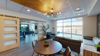 Located at 1609 SW Chandler Ave in Bend, Oregon, this single-floor community amenity space combines modern design with a welcoming, multifunctional layout. The space is organized into several distinct zones that flow seamlessly into one another. A spacious lounge area anchors the layout, featuring teal sectional sofas, a linear fireplace, and a warm wood-paneled ceiling with contemporary sputnik-style pendant lighting, creating a relaxed yet stylish atmosphere. Adjacent to the lounge, a game room accessed through a sliding barn door houses a foosball table and additional recreation space, accented by rich green walls and dark hardwood-style flooring throughout. A dedicated seating area features a round wood dining table and leather chairs beneath a statement chandelier, positioned near floor-to-ceiling windows that frame expansive outdoor views of the surrounding Central Oregon landscape. A live-edge wood communal table with industrial metal chairs provides an ideal setting for collaborative gatherings, also benefiting from generous natural light. The kitchenette is outfitted with wood-grain cabinetry, granite countertops, a penny-tile backsplash, and a sink, microwave, and coffee station. Nature-inspired artwork displayed throughout the space reinforces the Pacific Northwest aesthetic, completing this well-appointed and versatile community hub.