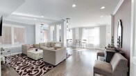 Located at 333 Central Avenue, this single-floor space offers an open, airy layout defined by a refined contemporary aesthetic. The main living area features light hardwood flooring, crisp white ceilings with recessed lighting, and elegant pendant light fixtures that serve as focal points throughout the space. Decorative columns subtly divide areas while preserving the open flow of the floor plan.

The lounge area is anchored by plush, neutral-toned seating arrangements set atop a bold patterned area rug, creating a warm and inviting atmosphere. A tufted sofa and coordinating sectional provide ample seating, complemented by a sleek, textured coffee table. Toward the rear of the space, a bar-height counter with tall seating offers a casual dining or gathering option.

Large windows dressed in sheer white drapery allow generous natural light to fill the space, enhancing the bright, neutral color palette of soft whites and warm grays. The overall ambiance is polished and welcoming, combining modern design sensibility with comfortable, functional furnishings suited for both relaxation and social gatherings.